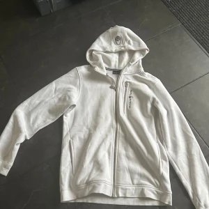 Xs/164 Vit hoodie från Sail Racing  - Säljer en snygg vit hoodie från Sail Racing med dragkedja framtill och på bröstfickan. Tröjan har huva med snörning och logga broderad på både bröstet och huvan. Perfekt för chill dagar eller när du vill ha en clean look.