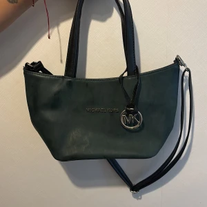 Grön handväska Michael Kors - Snygg grön handväska från Michael Kors med svarta handtag och axelrem. Väskan har ett stilrent MK-emblem i metall framtill och insidan är fodrad med logomönster samt har flera fack med dragkedja. Perfekt storlek för det viktigaste och en cool detalj till din outfit.
