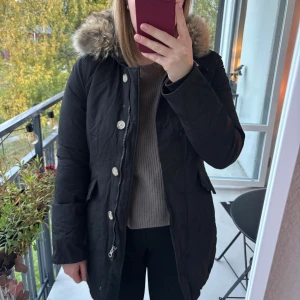 Vinterjacka Woolrich (Arctic parka) - Svart jacka med pälskrage och knappar. Jackan har fickor fram. Väldigt fint skick de då enbart är använd en säsong (resterande säsonger har de legat vakuumpackad, därför ser den tyvärr lite skrynklig ut men den hänger ut sig). Säljer pga för liten för mig! Ordinarie pris: 10500!