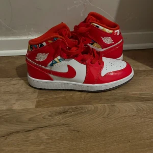 Nike Air Jordan 1 Mid röd/vit - Säljer ett par Nike Air Jordan 1 Mid i rött och vitt med färgglada detaljer runt ankeln. Skorna har klassisk snörning, rund tå och platt sula. Perfekta för dig som vill sticka ut med en ikonisk sneaker med coola färginslag.