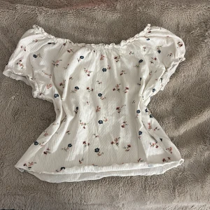 Vit blommig offshoulder croppad topp - Superfin vit croppad topp med blommigt mönster i rosa och blått från lager 157. Offshoulder-modell med korta puffärmar och rynkad kant upptill. Tunn, lätt och skön bomullskänsla, perfekt för varma dagar. Söt och trendig stil som lyfter din outfit.