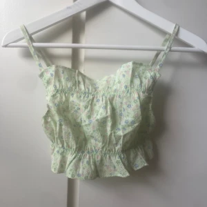 Blommig croppad linnetopp från H&M  - Supersöt croppad linnetopp från H&M i storlek XS. Toppen är ljusgrön med blommigt mönster i blått, rosa och vitt. Den har justerbara smala axelband och volangkant nedtill☺️