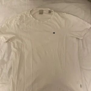 Vit t-shirt med symaskinslogga - Snygg vit t-shirt från Scotch & Soda i storlek M. T-shirten har rund hals, korta ärmar och en liten svart symaskinslogga broderad på bröstet. Tillverkad i mjuk bomull för skön känsla och enkel stil.