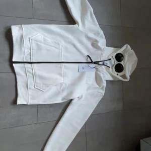 Vit hoodiejacka från C.P. Company - 🔥 C.P. Company Goggle Hoodie 🔥 Lyxig vit hoodie med den ikoniska huvan med glasögon 🕶️ och broderad logga 🤍 Mjuk, bekväm och perfekt för både vardag och streetwear-stil 💯 Exklusiv italiensk design 🇮🇹 – ett måste för dig som vill ha stil, kvalitet och något unikt ✨