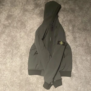Jacka från Stone Island - Säljer en jacka från Stone Island med huva och klassisk patch på vänster ärm. Jackan har dragkedja framtill, ribbade muddar och en clean, stilren look. Perfekt för dig som gillar streetwear och vill ha något som sticker ut. Storlek 156. Pris kan diskuteras 