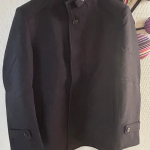 Svart ullrock  - Snygg svart rock med stilren ståkrage och knappar framtill. Jackan har en rak passform och detaljer med knappar vid ärmsluten. Perfekt för dig som gillar en clean och klassisk look. Materialet känns tjockt och varmt, idealiskt för kyliga dagar.