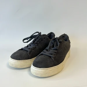 Mörkgrå mocka sneakers Selected Homme - Snygga mörkgrå sneakers från Selected Homme i mjuk mocka med vita platta sulor. Klassisk rund tå och snörning framtill. Perfekt för dig som gillar stilrena och enkla skor med en modern touch. Passar till jeans och hoodie eller en mer dressad look.