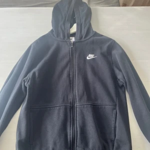 Svart Nike hoodie med dragkedja XL - Svart hoodie från Nike i storlek XL med vit logga på bröstet. Tröjan har huva, hel dragkedja framtill och två fickor. Tillverkad i mjukt material, perfekt för chill eller träning. Klassisk och enkel design som alltid funkar.