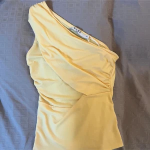 Gul oneshoulder topp från NA-KD - Säljer en ljusgul oneshoulder topp från NA-KD med snygga draperingar och tight passform. Toppen är ärmlös och har en asymmetrisk skärning som ger en cool och modern vibe. Perfekt att styla med jeans eller kjol för en trendig look. ( inte min bild)
