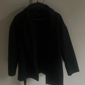 Svart kappa från Missay med bälte - Svart kappa från Missay med klassisk krage och bälte i midjan. Jackan har lång ärm och en stilren, rak passform som ger en snygg siluett. Perfekt för dig som gillar minimalistisk och tidlös stil.