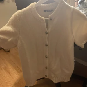 Vit kofta med kort ärm från Gina Tricot - Superfin vit kofta från Gina Tricot med korta ärmar och rund hals. Koftan har snygga silvriga knappar framtill och är stickad i ett mjukt material. Perfekt att slänga över en topp för en topp. Knappt använd😊