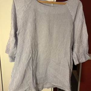 Randig blus från Rosebud i bomull strl: M - Säljer en fräsch blus från Rosebud i storlek M. Blusen är vit med tunna blå ränder, har trekvartsärm och är tillverkad i 100% bomull. Den har en loose passform och rund halsringning, perfekt för en chill och stilren look.