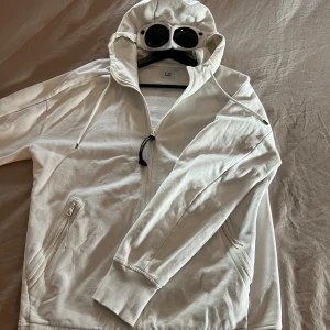 Vit hoodiejacka med goggles från C.P. Company - Vit full zip up hoodie från C.P. Company med ikoniska goggles i huvan. Jackan har dragkedja framtill, snörning i huvan och två sidofickor med dragkedja. Tillverkad i mjuk bomull för skön komfort och streetwear-vibe. Perfekt för dig som gillar unik design och detaljer.