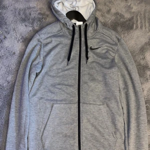 Grå Nike hoodie med dragkedja - Nike hoodie i ljusgrått med svart logga på bröstet, hel dragkedja och svarta snören i huvan. Tillverkad i mjuk Dri-Fit polyesterblandning, perfekt för chill eller träning. Fickor framtill och klassisk passform.
