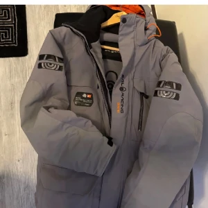 Sail Racing grå dunjacka med huva - Snygg och varm Sail Racing-jacka i toppskick, använd endast en vinter. Inga skador eller fläckar – riktigt fräsch! Flera fickor, rejäla dragkedjor och snygga logotyper på ärmar och bröst. Insidan är orange och jackan håller värmen perfekt. Nypris ca 8000 kr – nu endast 2250 kr! Unik modell som inte längre finns att köpa! Jackan är stor i storleken alltså large passar även xl 