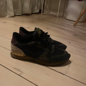 Valentino Garavani Rockrunner sneakers - Säljer ett par Valentino Garavani Rockrunner sneakers i mörkblått och brunt med svarta snören. Skorna har detaljer i mocka och skinn, samt en beige och vinröd häl med guldtonad plattform. Klassisk rund tå och platt sula. Perfekt för dig som gillar exklusiva streetwear-vibes. Litet hål på sulan som ni ser på sista sidan.