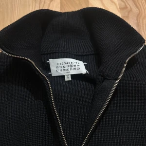 Svart stickad kofta från Maison Margiela - Svart stickad kofta från Maison Margiela med dragkedja framtill och klassisk ribbad struktur. Långärmad modell med hög krage och de ikoniska fyra vita sömmarna på ryggen. Perfekt för dig som gillar minimalistisk och stilren design.