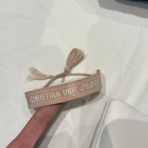 Beige armband från Christian Dior - Snyggt beige armband från Christian Dior med texten 'J'ADIOR' broderad i vitt. Armbandet har tofsar i ändarna och justerbar knytning för perfekt passform. Perfekt accessoar för att lyfta din outfit med en lyxig touch.