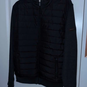 Svart quiltad pufferjacka från Lindbergh - Svart pufferjacka från Lindbergh med quiltad front och släta ärmar. Jackan har hel dragkedja, två fickor med dragkedja och diskret logga på ärmen. Perfekt för dig som gillar stilren och enkel design.