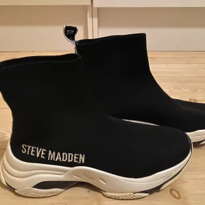 Svarta sock-sneakers från Steve Madden - Svarta sock-sneakers från Steve Madden med vit tjock sula och broderad logga på sidan. Skorna har en stickad ovandel som sitter tight runt foten och en cool dragflik med text bak. Perfekt för dig som gillar chunky sneakers med modern vibe.