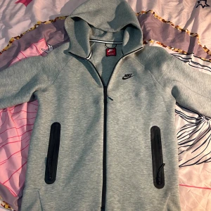 Grå Nike hoodie med dragkedja M - Snygg grå hoodie från Nike i storlek M. Tröjan har hel dragkedja framtill, två svarta fickor med dragkedja och klassisk Nike-logga på bröstet. Mjuk bomullsblandning och huva för extra komfort. Perfekt för chill eller träning.