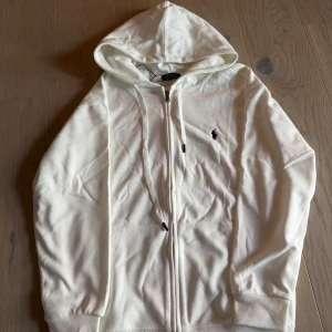 Vit hoodie från Polo Ralph Lauren - Snygg vit hoodie från Polo Ralph Lauren med klassisk logga broderad på bröstet. Tröjan har huva med snörning, dragkedja framtill och två fickor. Tillverkad i mjuk bomull som är skön att ha på sig. Perfekt för chill dagar och streetstyle.