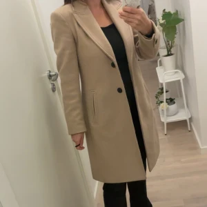 Beige kappa med knappar - Stilren beige kappa med klassisk krage och två svarta knappar framtill. Långärmad modell med raka linjer och diskreta fickor på sidorna. Perfekt för dig som gillar en clean och tidlös look.