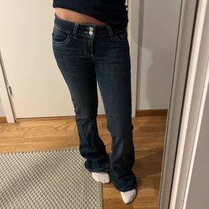 Mörkblå bootcut jeans från Nelly - Mörkblå jeans från Nelly med bootcut passform och låg midja. Snygga kontrastsömmar och dekorativ vit brodyr på bakfickorna. Jeansen har dubbla knappar framtill och klassisk femficksdesign. 