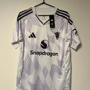 Manchester United bortatröja  - Säljer en Manchester United bortatröja från Adidas i vitt med ljuslila grafiskt mönster. Storlek M. Tröjan har korta ärmar. Materialet är lätt och andas, perfekt för match eller träning.