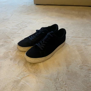 Svarta sneakers med vit sula - Snygga svarta sneakers med platt vit sula och matchande blåa snören. Perfekt för dig som gillar enkel och klassisk streetstil. Storlek 45, liten skada vid tårna på ena sidan förutom det är dom i väldigt bra skick. Meddela mig vid frågor eller intresse av fler bilder.