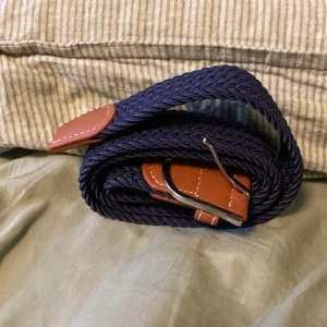 Mörkblå flätat bälte med skinn - Snyggt mörkblått flätat bälte med bruna skinn-detaljer och silvrigt spänne. Bältet har en flexibel och vävd design som gör det enkelt att justera. Perfekt för att ge din outfit en cool och stilren touch. Passar till jeans, chinos eller shorts.