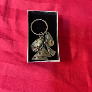 Nyckelring med egyptiska motiv och röda stenar - Cool nyckelring med antikguldiga berlocker i form av egyptiska motiv som farao och pyramid, dekorerade med små röda stenar. Kommer i en snygg silverfärgad presentask med rosett. Perfekt accessoar för dig som gillar unika detaljer.