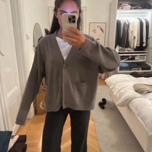 Grå kofta från H&M med fickor - Mysig grå kofta från H&M i oversized modell med två stora fickor framtill och knäppning med knappar. Koftan är stickad och har långa ärmar samt ribbade muddar vid ärmslut och nederkant. Perfekt för lager-på-lager och chill dagar.