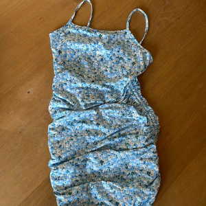 Blommig blå kortklänning med smala axelband - Supersöt kortklänning i ljusblått med små vita och gula blommor. Klänningen har smala axelband, rynkade detaljer på sidorna och en figurnära passform. 