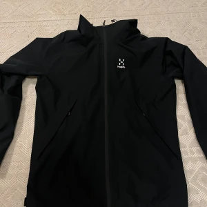 Svart vindjacka från Haglöfs med huva - Svart vindjacka från Haglöfs med minimalistisk design, Knappt använd. Liten defekt på en av fickorna (syns på sista bilden) 