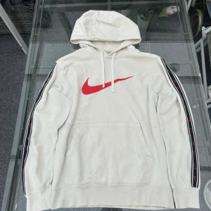 Vintage Nike hoodie vit med röd swoosh och loggor på ärmarna (S) - Superfin vintage Nike hoodie i färgen vit med röd swoosh-logga på bröstet och band med små swooshar längs ärmarna. Mjuk och bekväm insida i fleece, perfekt för både vardag, skolan eller streetwear-looken. Äkta Nike, tillverkad i Kambodja. I mycket bra skick, inga fläckar eller hål.  📏 Storlek: S (passar även XS för en lite oversized fit) 🧵 Material: Bomull & polyester 🎨 Färg: Vit med röd detalj  Perfekt för dig som gillar vintage Nike, Y2K, sportig stil eller casual outfits. 🔥 