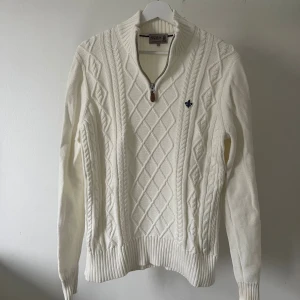  stickad half zip-tröja  - Snygg vit stickad tröja med half zip och hög krage från Morris . Tröjan har ett klassiskt kabelstickat mönster framtill och en liten blå brodyr på bröstet. Ribbstickade muddar vid ärmslut och nederkant ger en clean look. Perfekt för lager-på-lager under kyliga höst dagar. Modellen är 180cm. Noter en liten missfärgning k nacken. 