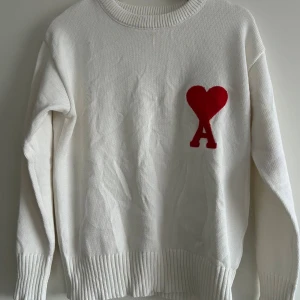 Vit stickad tröja från Ami Paris - Vit stickad tröja från Ami Paris med röd hjärtlogga och bokstaven A på bröstet. Tröjan har rund halsringning, ribbade muddar och lång ärm. Perfekt för dig som gillar stilrena plagg med en twist.