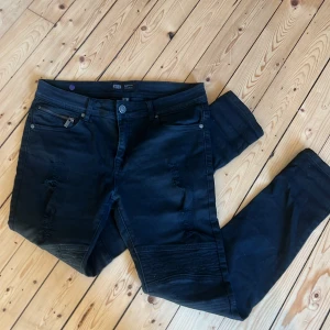 Svarta jeansbyxor från FSBN, 31/30 - Svarta jeansbyxor från FSBN med slitna detaljer och dragkedja på fickan. Byxorna har raka ben och coola sömmar vid knäna för en extra edge. Klassisk femficksmodell och bälteshällor. Perfekta för dig som gillar streetstil.