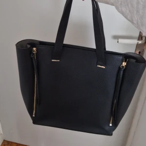 Svart axelväska med dragkedjor - Säljer en stilren svart axelväska i skinnimitation med två långa handtag. Väskan har snygga guldfärgade dragkedjor på sidorna som kan öppnas för att ge mer utrymme. Perfekt för dig som vill ha en rymlig och trendig väska till vardagen. Köpt på en väskbutik. Helt ny