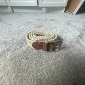 Beige flätat bälte  - Snyggt beige flätat bälte med bruna skinn-detaljer vid spännet och ändarna. Bältet har ett silvrigt metallspänne och passar perfekt till jeans eller chinos. Den flätade designen ger en avslappnad men stilren känsla.