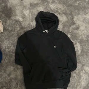 Svart hoodie från Lacoste med huva - Svart hoodie från Lacoste med klassisk krokodillogga på bröstet. Tröjan har huva med snörning, ribbade muddar och en stor magficka. Materialet är mjukt och känns som bomull, perfekt för chill dagar. Snygg och enkel design som funkar till allt.