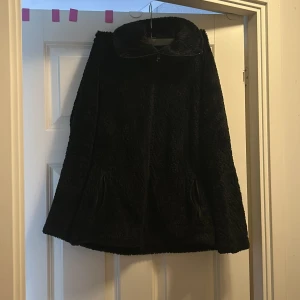 Svart teddyjacka från Crivit XL - Mysig svart teddyjacka från Crivit i storlek XL. Jackan har fluffigt material, hög krage och dragkedja framtill. Perfekt för kyliga dagar och har två fickor på sidorna. Enkel och stilren design som passar till det mesta.