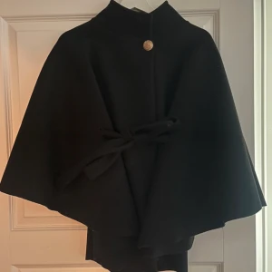 Svart cape med knytband och knapp - Säljer en svart cape med hög krage, dekorativ guldig knapp och knytband i midjan. Jackan har en stilren och svepande form som ger en elegant vibe. Perfekt för dig som vill sticka ut med en klassisk look.