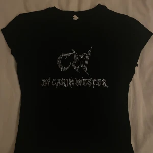 Svart topp från Carin Wester med strass - Säljer en svart kortärmad topp från Carin Wester med coolt CW-tryck och text i strass framtill. T-shirten har en klassisk rund halsringning och är gjord i mjukt material. Perfekt för dig som gillar statement-plagg med lite edge.
