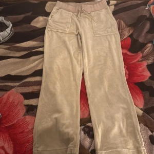 (Äkta)Beiga Juicy Couture mjukisbyxor - Snygga beiga mjukisbyxor från Juicy Couture i mjuk velour med bred resår och snörning i midjan. Byxorna har raka ben, stora fickor fram och bak med silverfärgade knappar och broderad JC-logga. Perfekta för en chill och trendig look.