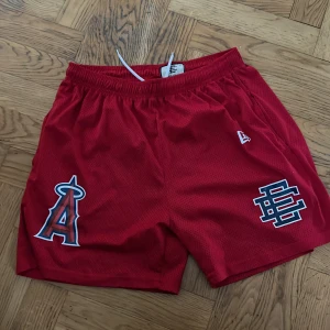 Eric Emmanuel shorts - Snygga röda eric emmanuel shorts med LA Angels-logga och coolt tryck på ena benet. Shortsen har elastisk midja med snörning och är perfekta för sport eller chill. Luftigt material som andas och ger en sportig vibe. Står lx men passar S/M