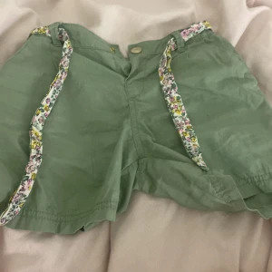 Gröna shorts med blommiga detaljer - Snygga gröna shorts med blommiga band längs sidorna. De har en avslappnad passform och stängs med knapp och dragkedja framtill. Perfekta för varma dagar när du vill ha en chill och färgglad look.