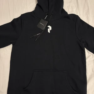 Svart hoodie från Peak Performance S - Svart hoodie från Peak Performance med stor vit logga på ryggen och mindre logga på bröstet. Klassisk känguruficka framtill, dragsko i huvan och mjukt material. Perfekt för chill eller streetstyle. Helt oanvänd med prislapp.