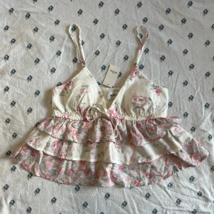 Blommig babydoll peplum volangtop från Hollister - Supersöt babydoll peplum blommig topp från Hollister med volanger och smala axelband. Toppen är vit med rosa och gröna blommor, har v-ringning, knyt fram och avtagbara inlägg. Perfekt för sommarens alla tillfällen!